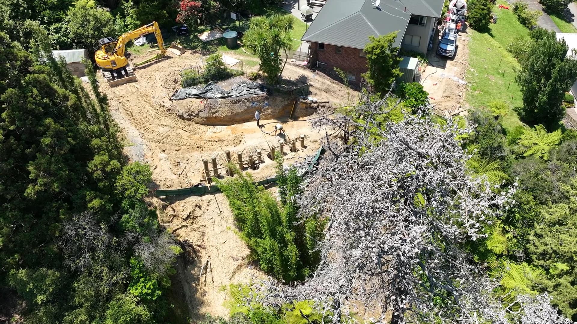 Aerial view of an Auckland hillside section under excavation, with an excavator, a cut bench and a line of retaining posts below a house