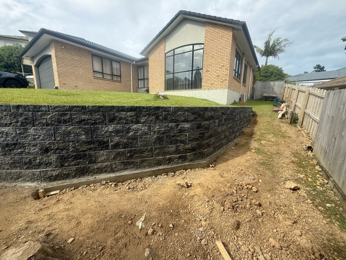 Curved dry-stack concrete block retaining wall holding a lawn above a levelled strip beside an Auckland house