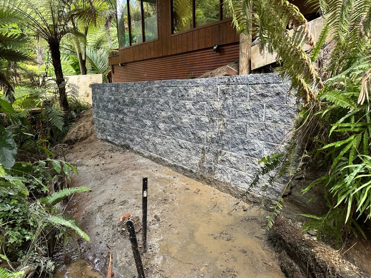 A dry-stack concrete block retaining wall holding ground beneath a timber-clad house, surrounded by tree ferns, with a wet excavated access below