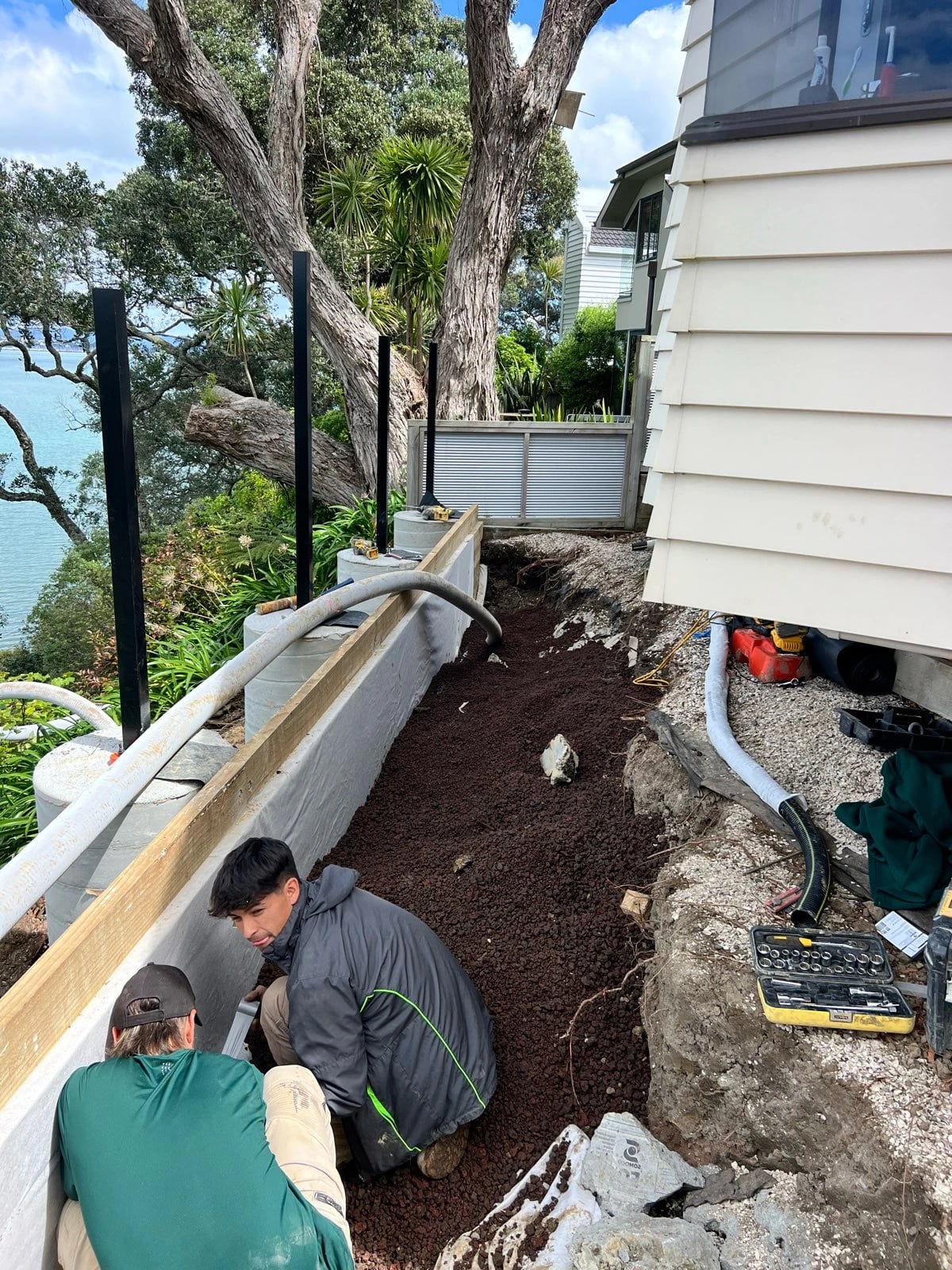 Two workers laying scoria drainage aggregate and flexible subsoil pipe behind a wall beside a house, with the sea behind them