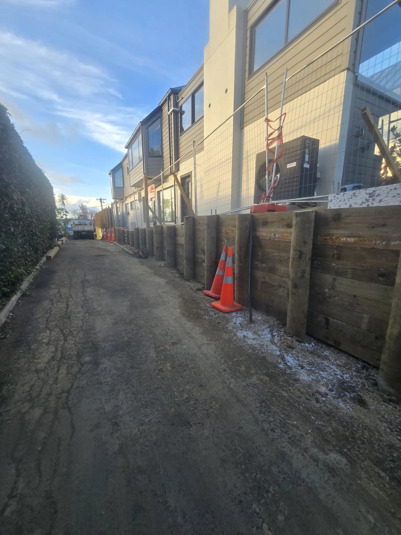A timber pole retaining wall running the length of a shared driveway beside townhouses, with traffic cones along its base