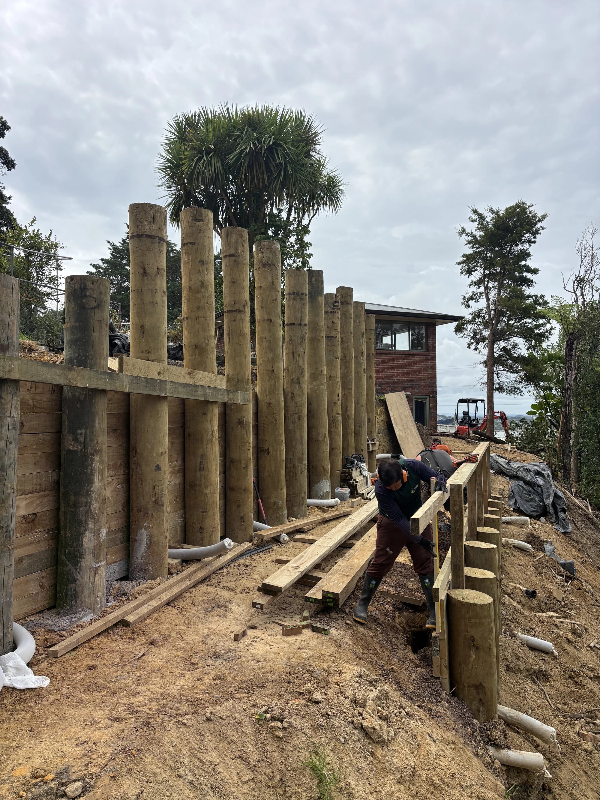 A worker in hearing protection lifting a timber rail into place along a pole retaining wall, with a house and the sea behind