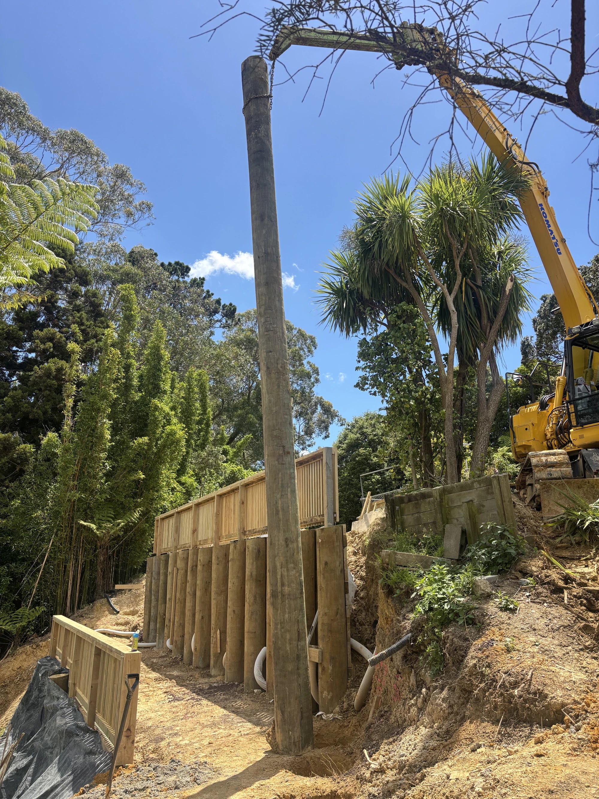 A treated timber pole slung from a long-reach excavator above a part-built retaining wall on a bush-clad Auckland slope