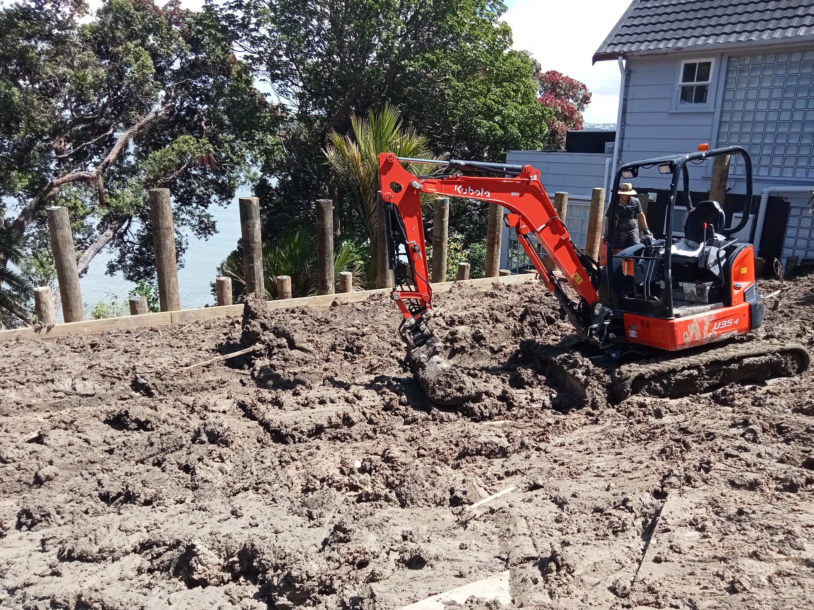 Row of timber poles set along a levelled bench on a coastal Auckland section, with a compact excavator working beside a house