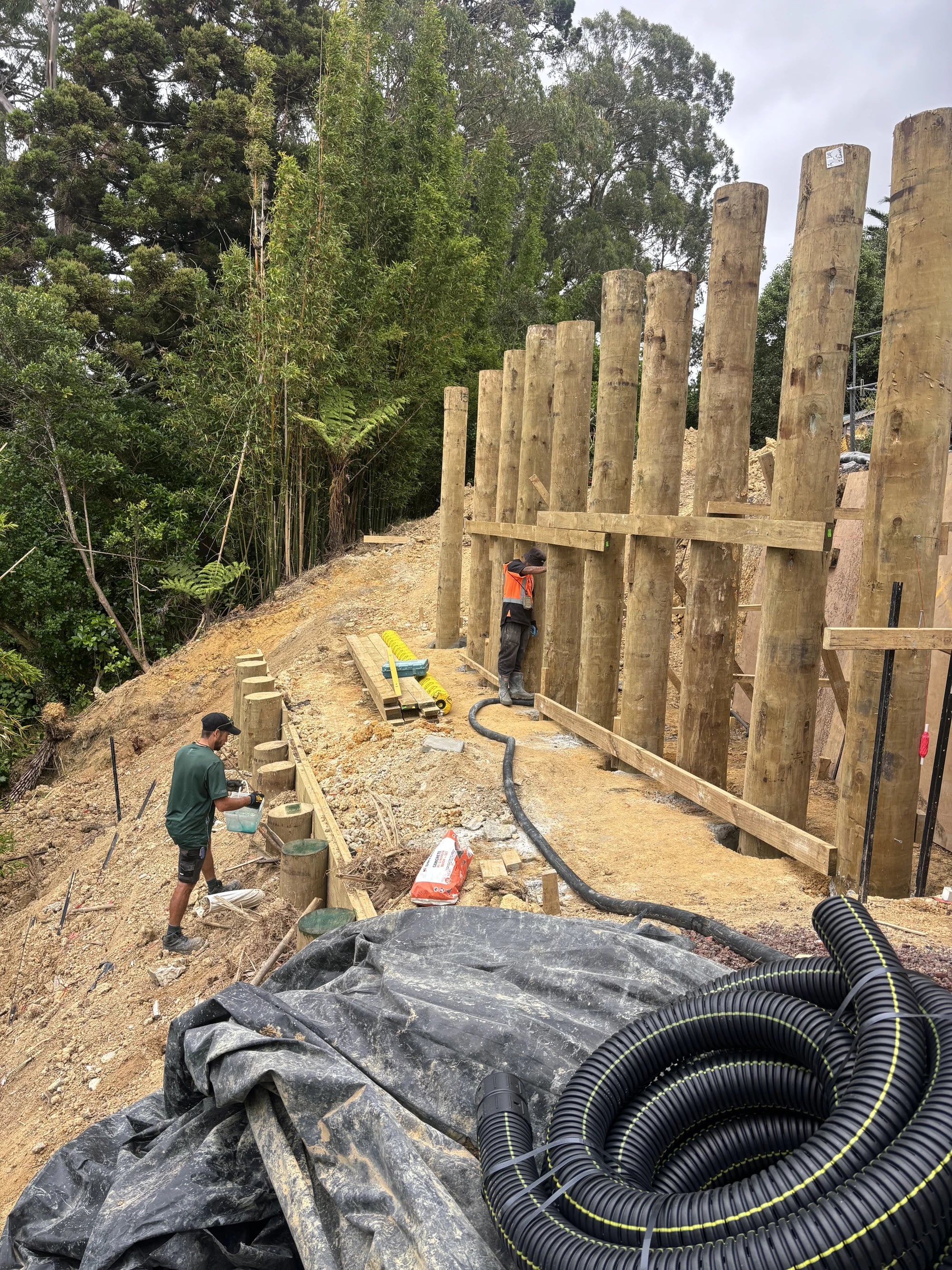 A timber pole retaining wall part-built along a steep bush slope, with two workers, timber walings and coiled drainage pipe on the bench
