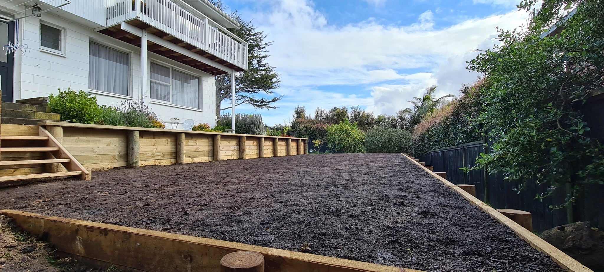 Level terrace of topsoil enclosed by low timber retaining walls and timber steps, beside a white Auckland house