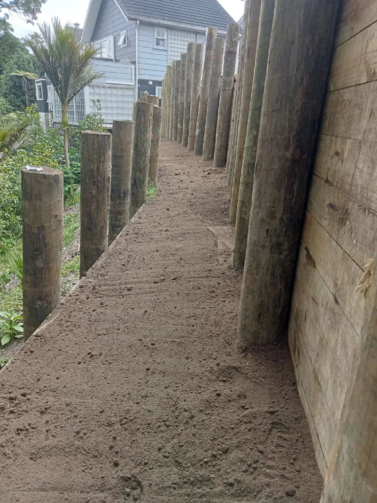 Tall timber pole retaining wall with horizontal lagging, running the length of a narrow bench beside an Auckland house