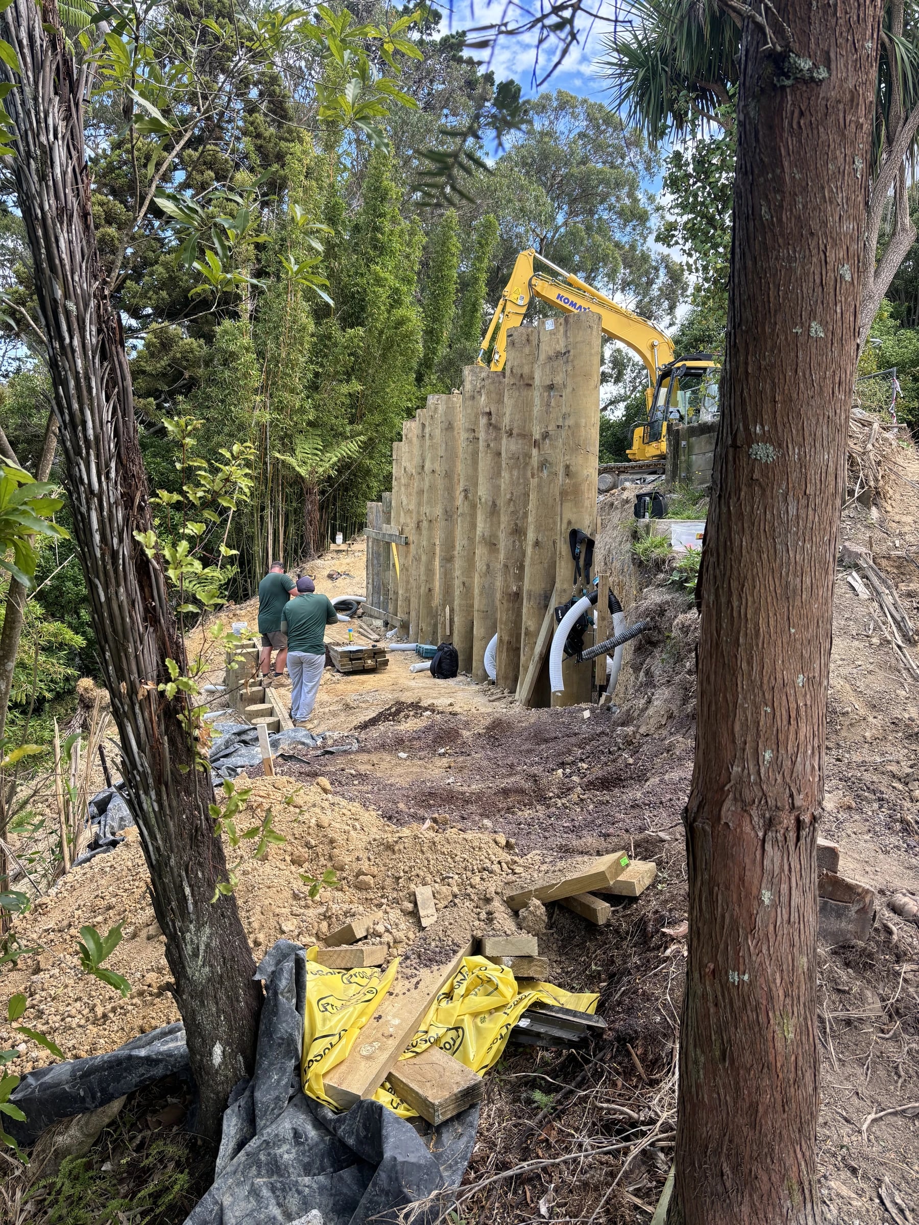 A tall timber pole retaining wall seen from below on a bush section, with an excavator on the ground above and two workers at its base
